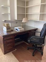 Wide view of wood executive desk, black leather office chair, two desk lamps, and corner built-in shelving