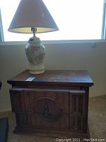 Nightstand with floral ceramic lamp placed on top, lamp turned on in front of window