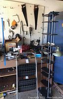 Various hand saws hung on a pegboard, yellow drill, gloves, vintage folding light, metal step ladder, shelving unit and other tools stacked on/around a workbench.