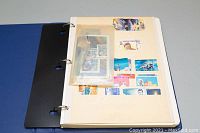 Open binder showing multiple stamps mounted on an album page with a small plastic pouch containing loose stamps.