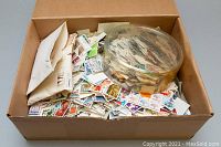 Large cardboard box filled with assorted loose Canadian stamps, some in small envelopes and some loose in a plastic container on top.
