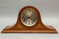 Front view of wooden mantel clock with curved top, showing silver clock face with black numerals and decorative inlaid wood accents.
