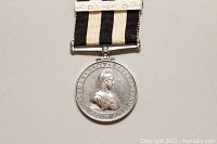Close-up of the silver-toned medal showing Queen Victoria's profile, round with Latin inscriptions, attached to a black and white striped ribbon with a decorated white bar.