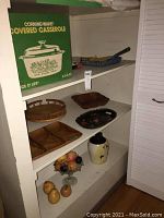 Photo shows a Corning Ware covered casserole in its original green and white box, vintage wooden trays, a dark wood bowl, a black lacquered tray, a vintage pottery cookie jar, and faux fruit on a glass footed plate.