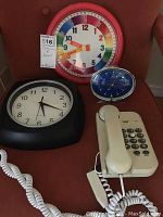 Photo showing two wall clocks, one colorful with multi-colored numbers, and one black clock with white face, alongside a blue mantel clock and beige corded phone on a red fabric surface.