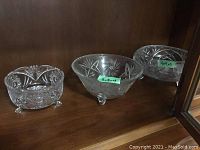 Six clear crystal and cut glass footed bowls with various decorative cut patterns including pinwheel and starburst designs, displayed on a wooden shelf.