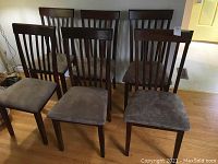 Six wood-framed dining chairs with dark wood finish and upholstered seats in soft brown fabric. They feature vertical slats on the backrests and show some wear including scratches.
