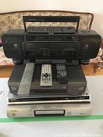 Stacked four electronics: Lloyds boombox with two detachable speakers on top, Toshiba VHS player beneath it, Konka DVD/VCD/CD player next, and Panasonic DVD player at bottom. Three remotes placed on top then stacked devices.