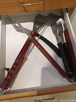 Drawer containing BBQ utensils including wooden handled lifter, forks, spatula, and black handled tongs