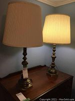 Pair of two antique brass trilight lamps on wooden table with cream pleated shades, one lamp turned on showing warm light.