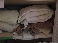 Shelves with folded linens including crocheted bedspreads and various sheets and pillowcases in white, beige, and pastel tones.
