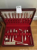 Closed wood case with lid open showing rows of silverplate flatware arranged in red velvet slots, including forks, knives, spoons, ladle, butter spreader, sugar tongs, and serving spoon.