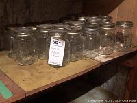 Nineteen small clear glass mason jars lined up on a wooden shelf, showing metal screw-on lids and embossed lettering on some jars.