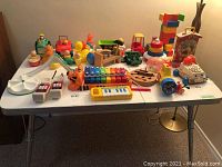 Overhead view of entire assortment of toddler toys on white folding table including light toys, musical instruments, push and pull toys, and stacking toys in various colors and styles.