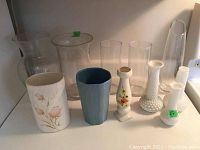 Front angled view showing assortment of glass, ceramic, and milk glass hobnail vases in various sizes and colors including white, clear, blue, and floral patterns.