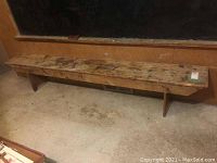 Full view of the long wooden bench showing top and supporting legs against a wall with concrete floor beneath.