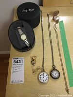 Overview of the watches, rings, and brooch on a wooden surface including the Citizen boxed wristwatch, 3 pocket watches, 2 rings, and 'Grace' brooch.