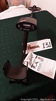 Metal detector showing full length, black with foam handle and armrest, lying on green fabric surface with lot number and instruction manual.