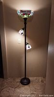 Full view of stained glass floor lamp turned on, showing the main top shade glowing with visible stained glass floral design and two smaller side shades lit.