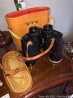 Binoculars placed with leather case and cover on a wooden surface.