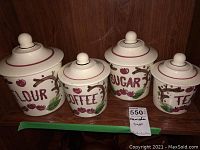All four ceramic canisters labeled Flour, Coffee, Sugar, and Tea with lids. Positioned on a wood shelf.