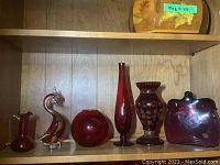 Full view of six cranberry glass items including vases and a purse vase arranged on wooden shelf.