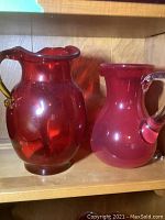 Two colored glass jugs, red with yellow handle and pink with clear handle on wooden shelf with visible damage on red jug handle.