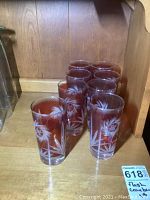 Eight cranberry flash glass tumblers with etched leaf and geometric designs arranged in two rows on a wooden shelf.