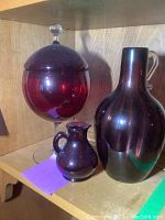 Full view showing three pieces of the glassware lot on a wooden shelf: large covered dish with pedestal, medium vase, small pitcher-style vase, all in deep red/purple glass.