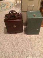 Photo showing the vinyl rolling briefcase next to green metal safe with key attached to latch, placed on carpeted floor.