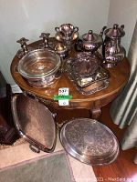 Full view of silver plate coffee and tea pots, candlesticks, trays, serving dish with glass lid on round wooden table