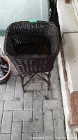Front angled view of the square wicker planter on a four-legged wicker stand placed on a paved surface with some leaves and debris inside.