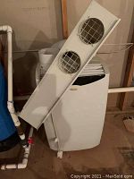 Front view of white Danby Premiere portable air conditioner with attached white vent cover featuring two circular grates, situated in a basement with piping nearby.