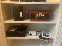 Full view of white shelving unit with black Vizio subwoofer, vintage-style record player on top shelf, Onkyo DVD changer/receiver with remote, white DB Power projector, assorted sheet music and piano lesson books on middle and bottom shelves
