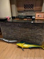 Two colorful metal fish wall art pieces lying on the floor in front of a bar with stone facade and wooden cabinetry with wine rack above.