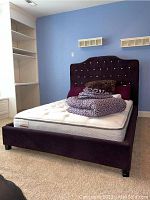 Front view of purple velvet bed frame with headboard, footboard, side rails, mattress and linens piled on top