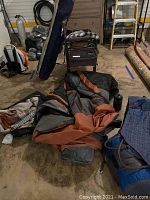 Photo showing a folded orange and gray tent or sleeping bag with various camping supplies around on a concrete floor.