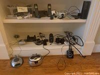 Shelf with assorted electronic devices including cordless phones, Amazon Echo speaker, ultrasonic cleaner, pencil sharpener, wireless transmitter, and computer speakers
