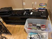 Wide view of electronics and media items on TV stand and floor, showing DVD players, receiver, cables, DVDs stacked, and a large bin of CDs.