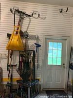 Wide view of garden tools hanging on a slat wall including snow shovels, metal garden rakes, broom, and other tools mounted vertically with door on right