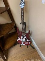 Full view of the red Ibanez electric guitar with white gear-shaped decals, AOL and CMPS Summer 2007 markings, positioned on a black guitar stand by a wall.