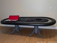 Full view of oval poker table with black padded bumper, custom grey felt top with skull graphic reading “Punisher’s Pub,” and double pedestal grey wooden legs.