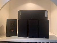 Front view of four smaller Polk Audio speakers and one larger Polk Audio speaker arranged on a shelf.