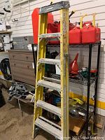 Yellow and silver 6-foot Husky metal stepladder standing upright near metal shelving with red gas cans on middle shelves