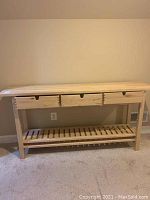 Full front view of the wooden workbench style table showing its three drawers and bottom slatted shelf against a beige wall and carpeted floor.