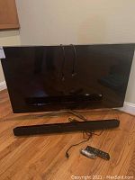 Front view of Samsung TV on stand with Vizio soundbar placed in front, power and connection cables draped over TV and floor, Samsung remote beside soundbar.