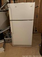 Full view of white Whirlpool top-freezer refrigerator in basement setting. Shows general condition and design.