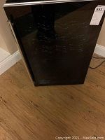 Front view of the black wine cooler with glass door and metal handle on wooden floor.