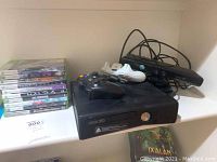 Front view of Xbox 360 console stacked with two controllers (black and white) and Kinect sensor on top, cables visible.