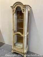 Front angled view of tall cream colored curio cabinet with glass front and sides, ornate carved top, showing built-in light inside and mirrored back panel with glass shelves.